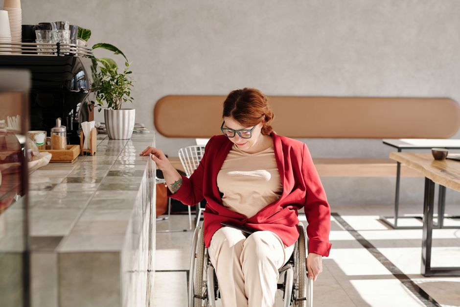 A woman in a wheelchair wearing a red blazer sits at a café counter, enjoying leisure time indoors.
